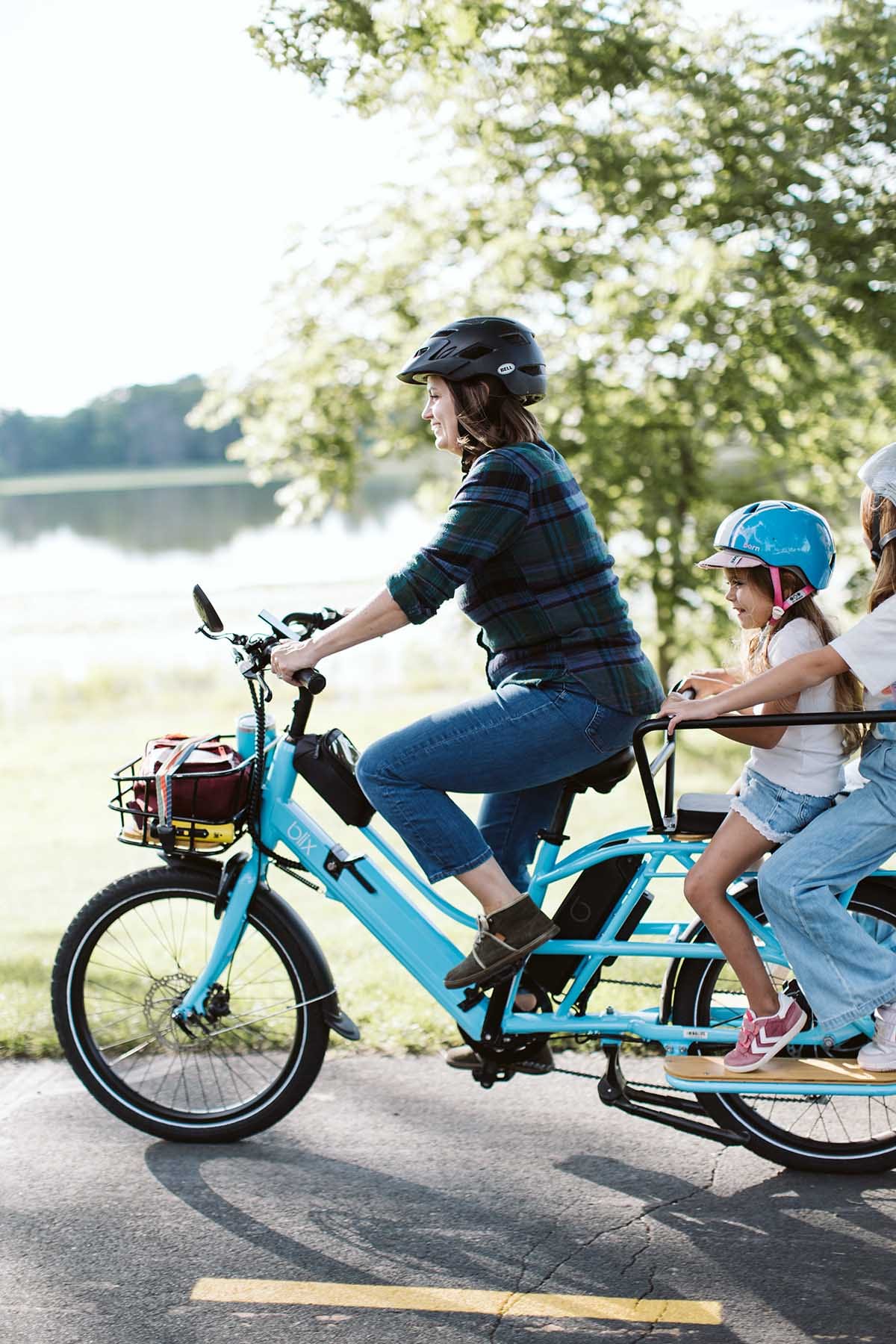 A smiling mom rides her blue Blix cargo bike with two young girls on the back, heading to school by lake in Minneapolis, Minnesota. The bike has a hidden BikeFinder tracker for theft protection.